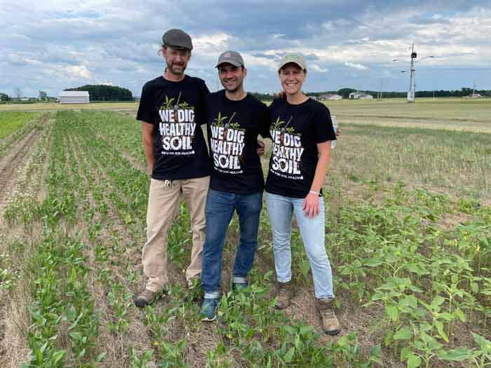 three people standing an agricultural field wearing black t-shirts with the phrase "We Dig Healthy Soil":