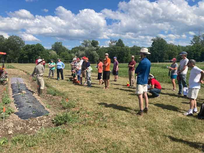 demonstration of using tarps for weed suppression at a soil health workshop. Participants are standing in an agricultural field, blue sky with clouds in background