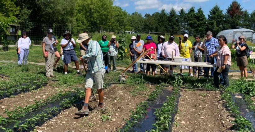 More than a dozen people gathered in an agricultural field with crops in the foreground and most people in back gathered near a soil health demonstration table.