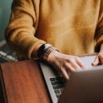 Person wearing a camel colored sweater typing on a laptop. Only hands and torso are in view.