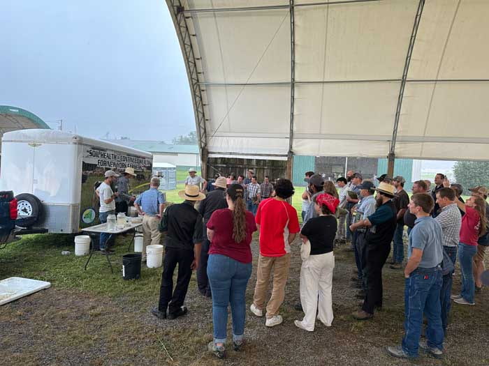 Large group of people gathered outdoors to watch soil health demonstrations at a table in front of the New York Soil Health trailer. 